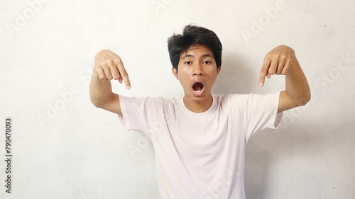 young asian man poses surprised while pointing down on white background