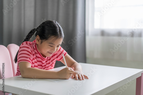 learn from home by asian child or kid girl student smile writing on paper to draw learning studying or young people fun doing homework in classroom homeschool on white table or desk and window light