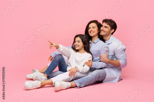 Excited european family of three pointing finger aside, posing sitting on pink studio background, girl showing and advertising