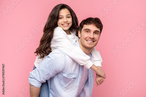 Cheerful father giving his pretty daughter piggyback ride, while they are both looking at camera and smiling over pink background