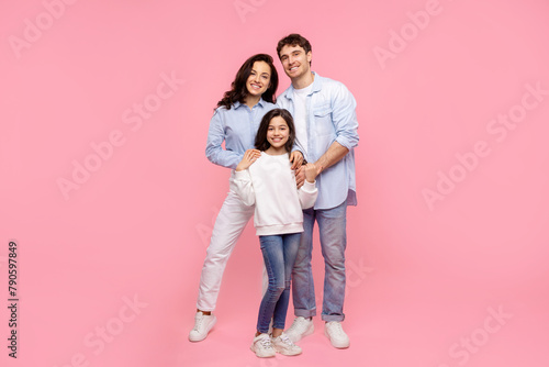 Happy mom dad with child daughter cuddling and smiling at camera, posing isolated on pastel pink background, full length shot