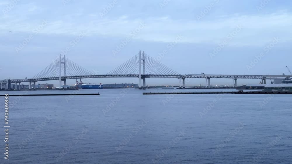 Distant view of a suspension bridge over serene water with boats, under a cloudy sky Distant view of a suspension bridge over serene water with boats, under a cloudy sky