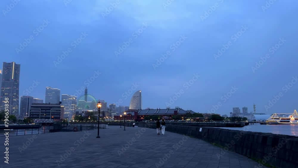 Dusk view of a cityscape with a couple walking along the waterfront, buildings lit up in the background Dusk view of a cityscape with a couple walking along the waterfront, buildings lit up in the background