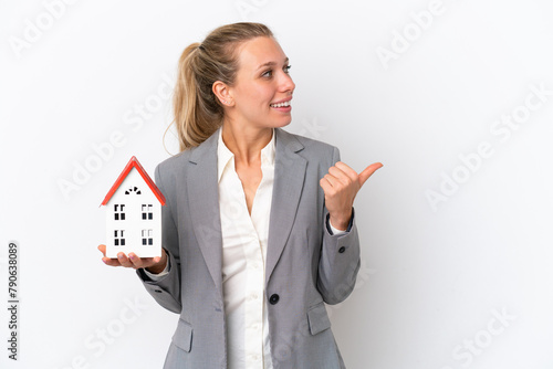 Real estate agent woman holding a toy house isolated on white background pointing to the side to present a product