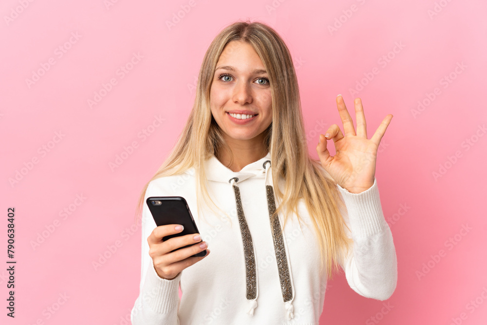 Young blonde woman isolated on pink background using mobile phone and doing OK sign Young blonde woman isolated on pink background using mobile phone and doing OK sign