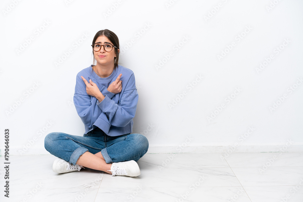 Young caucasian woman sitting on the floor isolated on white background pointing to the laterals having doubts Young caucasian woman sitting on the floor isolated on white background pointing to the laterals having doubts