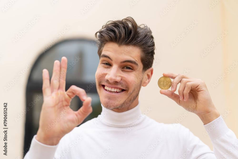 Young caucasian man holding a Bitcoin at outdoors showing ok sign with fingers Young caucasian man holding a Bitcoin at outdoors showing ok sign with fingers