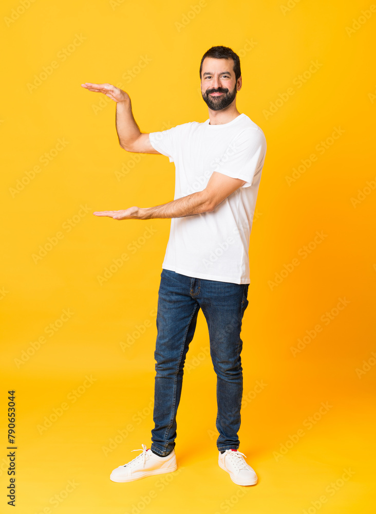 Full-length shot of man with beard over isolated yellow background holding copyspace to insert an ad