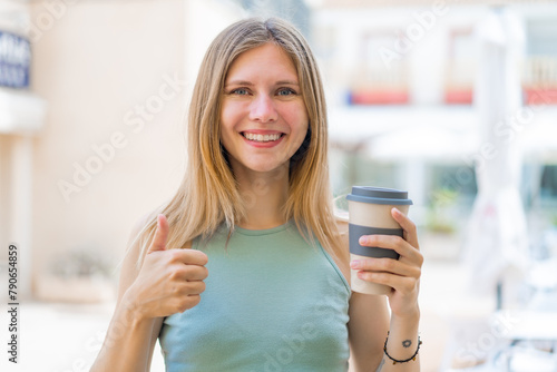 Young blonde woman holding a take away coffee at outdoors with thumbs up because something good has happened
