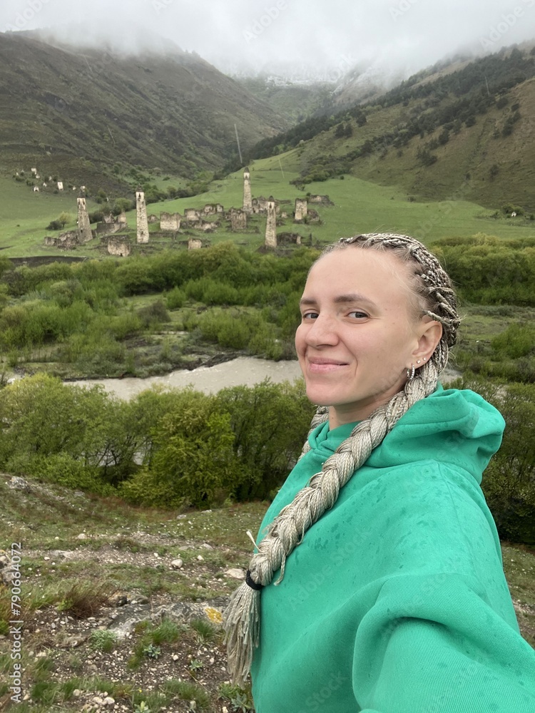 Naklejka premium A girl on the background of the medieval Targim tower complex in the Caucasus mountains, surrounded by greenery. The Caucasus Mountains on a cloudy day in Ingushetia. Russia