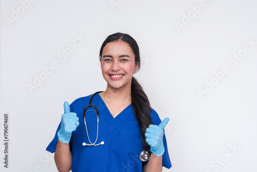 Smiling Asian female nurse in blue scrub suit giving thumbs up, isolated on white