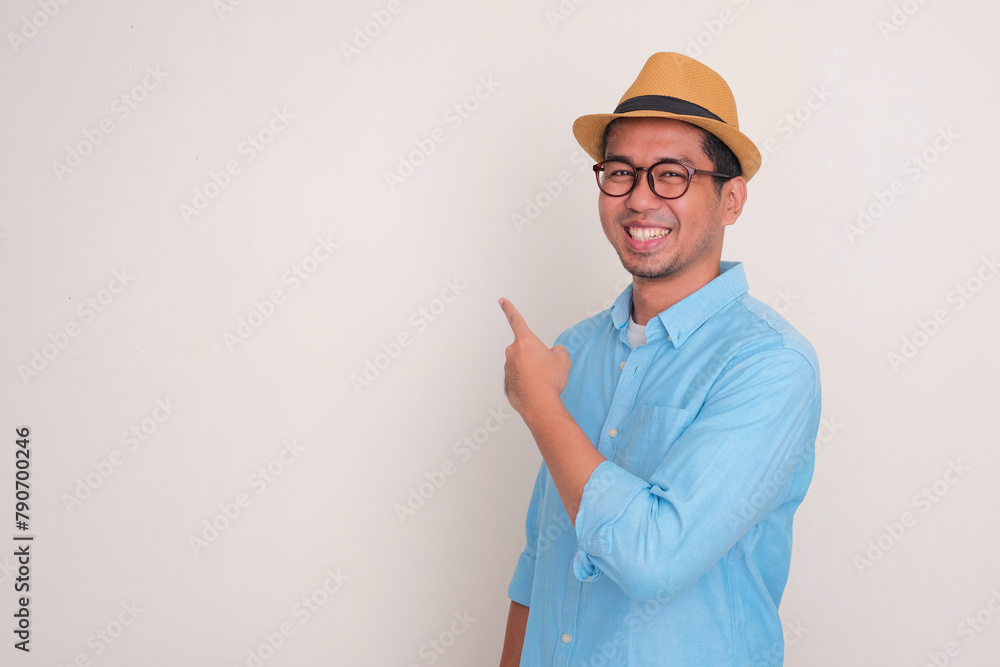 A man wearing beach hat smiling at the camera with one hand pointing behind him A man wearing beach hat smiling at the camera with one hand pointing behind him