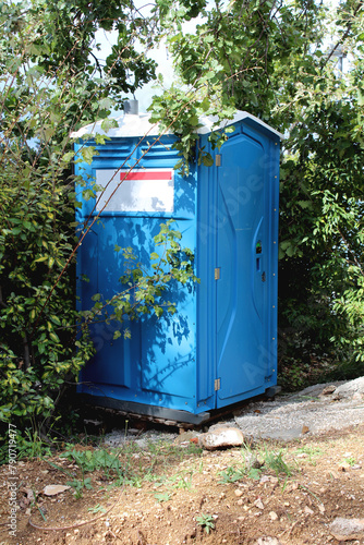 New portable unisex blue with white top ecological toilet or portable chemical toilet on wooden pallet at local construction site surrounded with dense trees