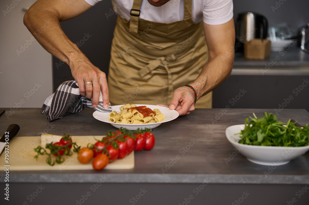 Close-up man in beige chef apron, using kitchen towel, removes traces of tomato sauce on the white plate with Italian pasta before serving the dish. Fresh organic ingredients on the kitchen countertop