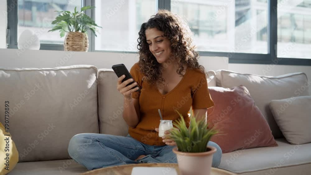 Video of beautiful woman eating healthy yogurt while using smartphone sitting on couch at home. Video of beautiful woman eating healthy yogurt while using smartphone sitting on couch at home.