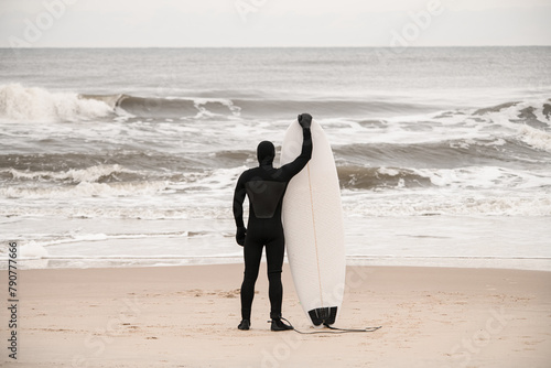 Slim male surfer in black wetsuit standing on the seashore