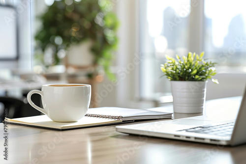 A white coffee cup sits on a table next to a laptop and a plant