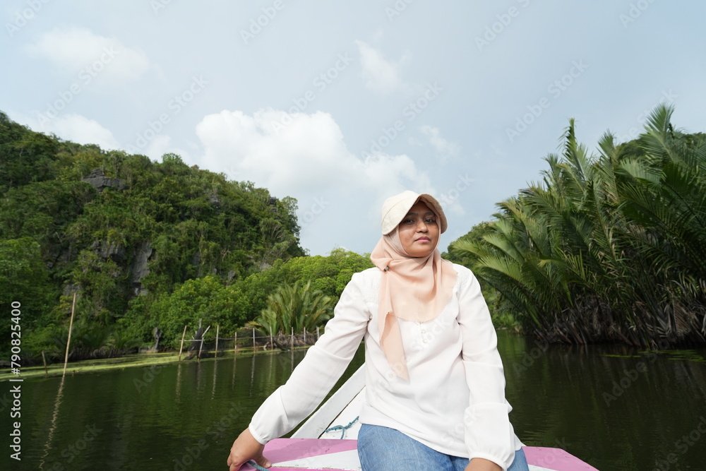 Fototapeta premium Portrait of woman sitting in wooden boat. Asian woman wearing a hijab sits on a wooden boat that is traveling along a river with mangrove trees around it