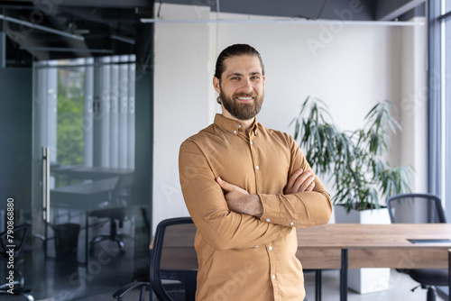 A cheerful, bearded businessman standing confidently with arms crossed in a contemporary office environment, exuding professionalism and positivity.