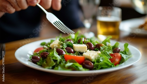 A person sits at a wooden table with a plate of fresh salad with cheese and olives, flanked by two cups. A spoon rests near the top of the plate, likely for serving.