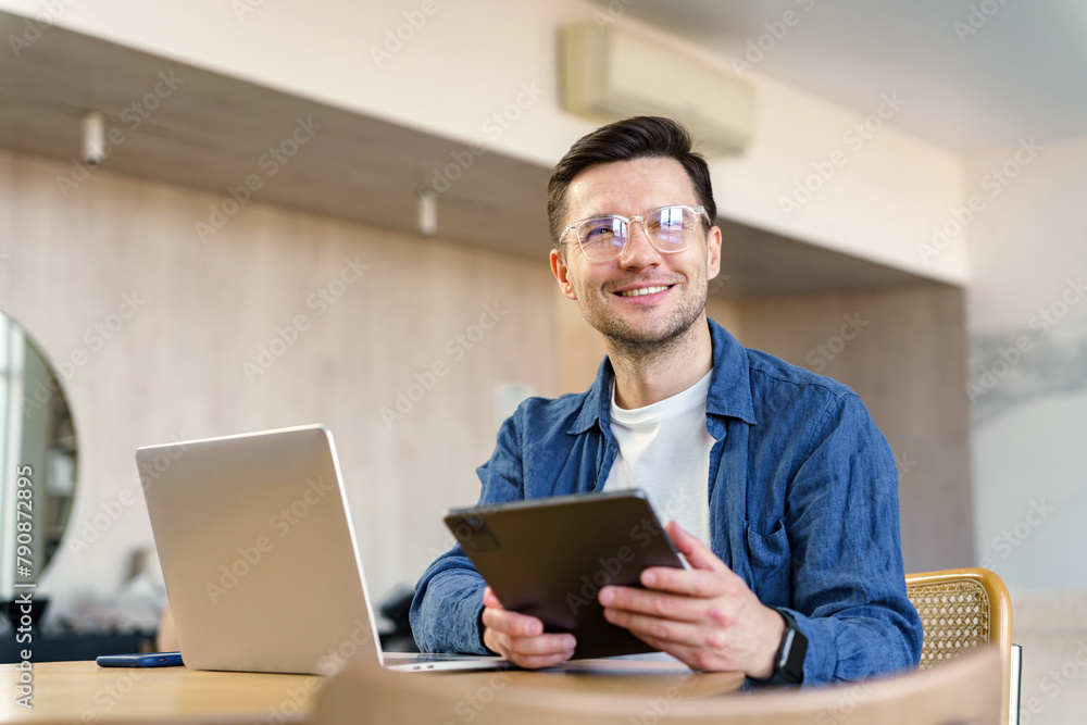 Charming professional smiles while using a digital tablet, seated with laptop at a chic workspace in a modern home. Charming professional smiles while using a digital tablet, seated with laptop at a chic workspace in a modern home.