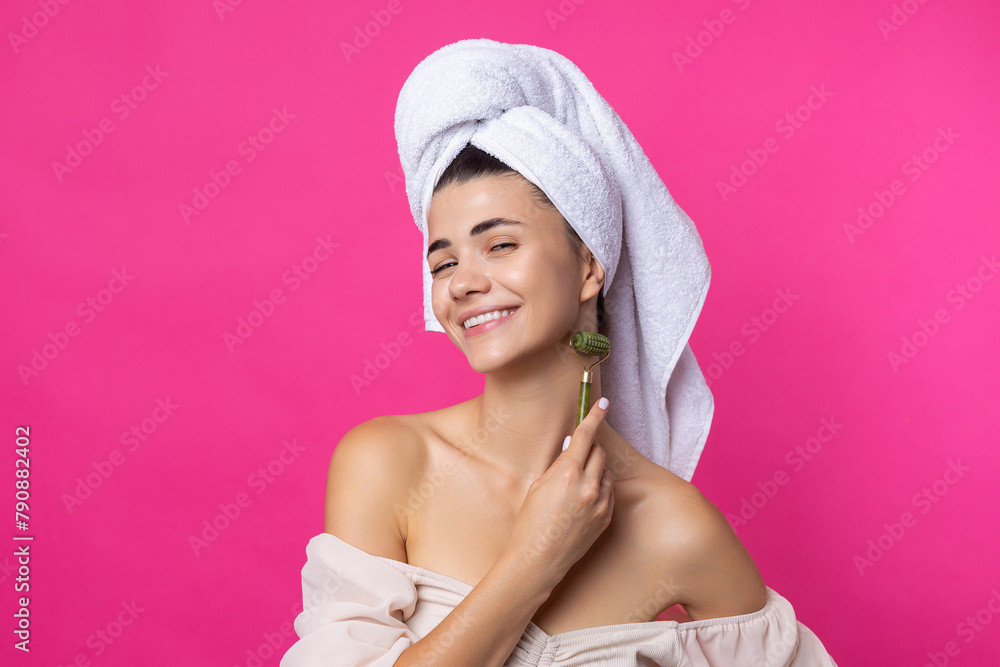 A beautiful cheerful attractive girl with a towel on her head holds a cosmetic roller near her face against a pink background. A beautiful cheerful attractive girl with a towel on her head holds a cosmetic roller near her face against a pink background.