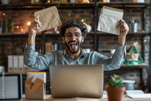 An excited Middle Eastern man in casual attire, sitting at an office desk with his laptop open and holding up documents to the camera, exuding happiness while celebrating success or winning happily.  photo