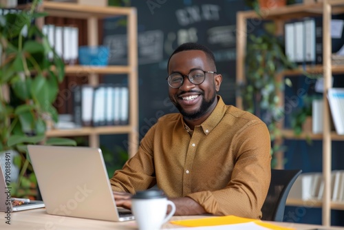 A smiling man in glasses is sitting at the table with laptop and writing on paper, working as an IT technician or project manager of web development team in modern office space. Man dressed light