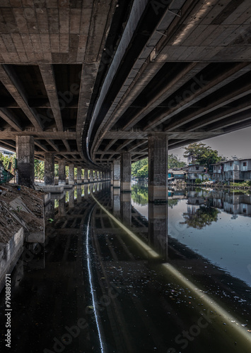 Perspective view of concrete pillars under Khlong Toei Expressway with calm surface water reflection and houses are old along the Khlong Phra Khanong. Space for text, Selective focus.