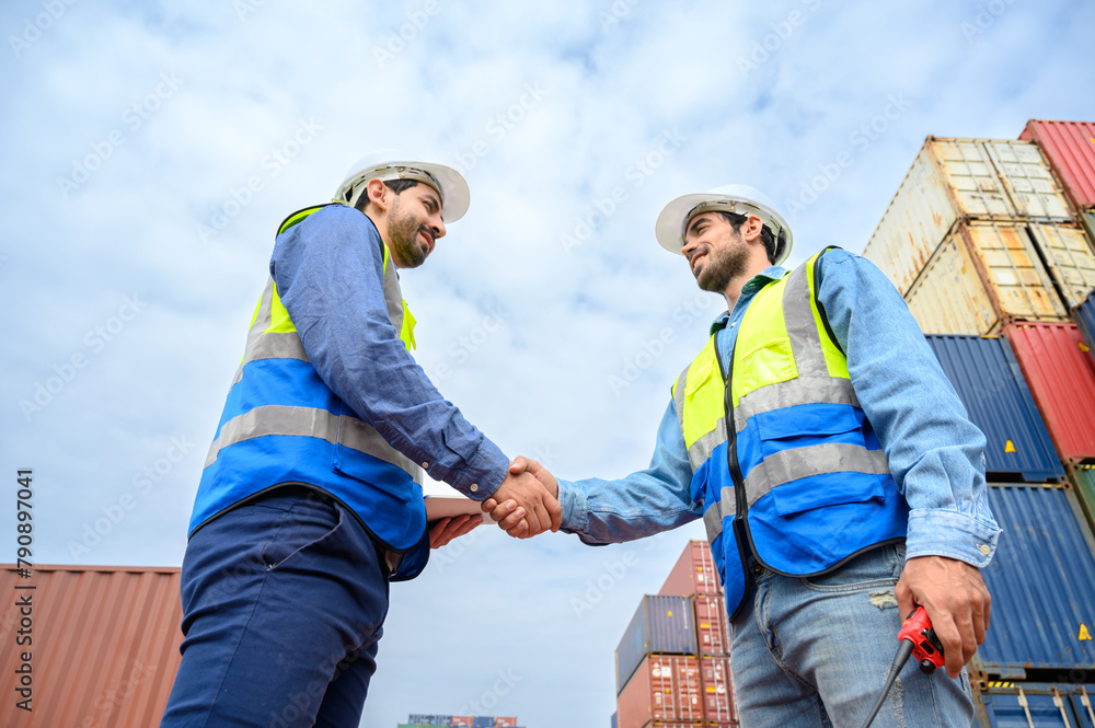 Two engineer container cargo wearing white hardhat and safety vests in successful handshake together at container yard. success in logistics business import export industry