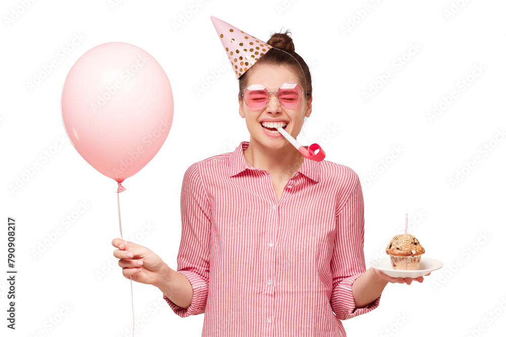 Obraz premium Young happy girl in glasses and birthday hat, blowing whistle, holding pink balloon and cupcake, laughing and enjoying party