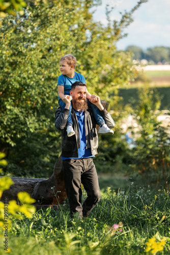 Dad and son walking in nature on a sunny summer day. The father carries his son on his shoulders, the boy is raised high and looks around and admires the summer nature