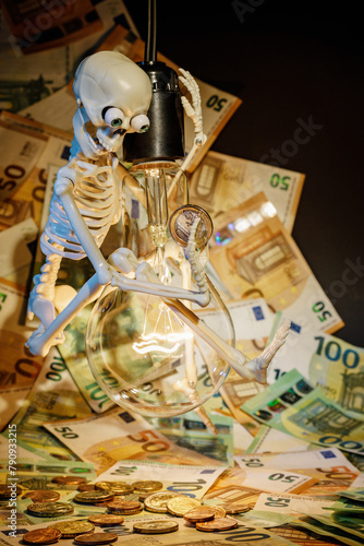 A toy skeleton with a one euro coin in his hand on a glowing light bulb on a background of euro banknotes. Concept Let's save electricity