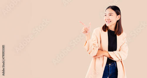 Happy, young woman in a chic beige coat pointing to the side with a joyful expression, isolated on a beige background.
