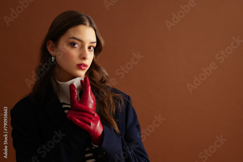 Fashionable confident woman wearing stylish red leather gloves, turtleneck sweater, classic blue woolen coat, posing on brown background. Studio fashion portrait. Copy, empty, blank space for text 