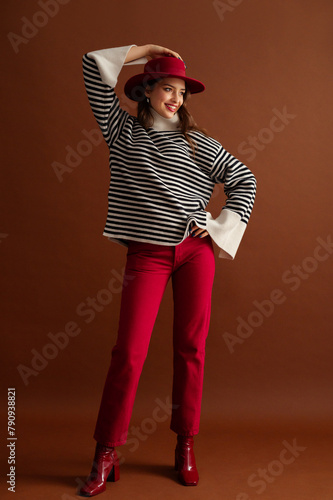 Fashionable happy smiling woman wearing stylish striped oversize turtleneck sweater, red jeans, hat, patent leather ankle boots, posing on brown background. Full-length studio fashion portrait