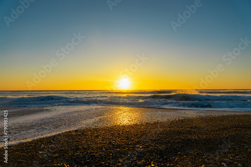 Au bord de l'océan Atlantique, dans la baie d'Audierne en Bretagne, un coucher de soleil teinte le ciel de nuances jaunes et bleues, offrant des reflets éblouissants sur le sable mouillé et les galets