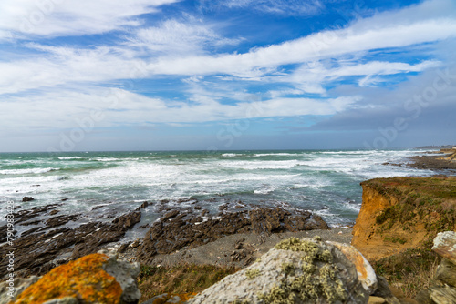 Sur le littoral atlantique de la baie d'Audierne, ciel bleu, nuages blancs, rochers lichens orange, sillons rocheux, écume de mer.