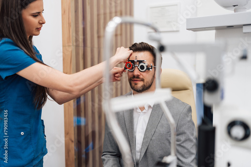 Young female doctor or oculist putting ophthalmic glasses or trial frame on a smiling handsome male patient. Modern clinic for diagnosis and treatment of eye and sight diseases. Eyecare concept.
