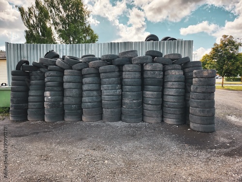 Neatly arranged pile of worn-out tires in various sizes stacked up in an organized manner outdoors on a vacant lot next to a sturdy chain link fence under a dramatic and overcast sky