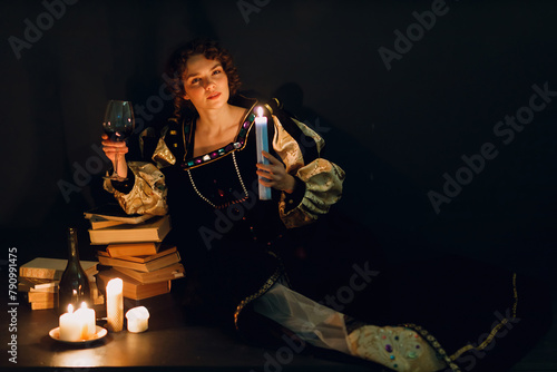 Young adult woman dressed in a medieval dress sitting near ancient books and candles with bottle of red wine and holding a glass of wine in darkness