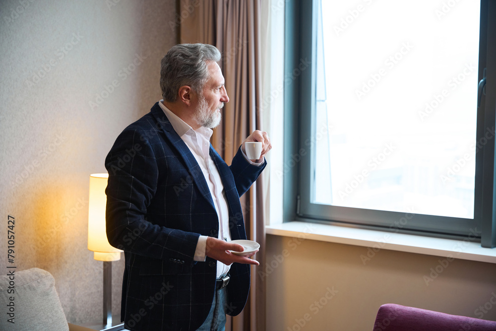 Fototapeta premium Caucasian businessman drinking tea or coffee from cup and looking from window