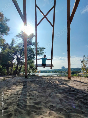 A young woman swings on a huge handmade wooden swing in an amusement park on the sand. Rear view