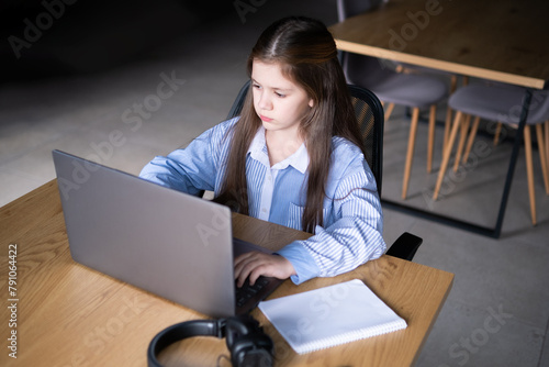 A cute small girl sitting alone at a table looking in a laptop screen doing her distance education hometask in a big room photo