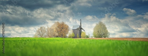 panoramic landscape with field of green wheat and old wooden mill under rainy clouds in spring day