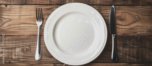 A white, vacant plate accompanied by utensils, set against a backdrop of wooden texture.