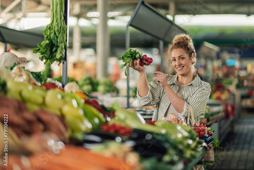 Young buyer with radish in hand staining near stall at food market.