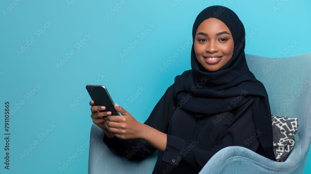 Happy African Muslim woman in abaya hijab using mobile phone while sitting on the armchair and looking at the camera on isolated blue background with space for copy Happy African Muslim woman in abaya hijab using mobile phone while sitting on the armchair and looking at the camera on isolated blue background with space for copy