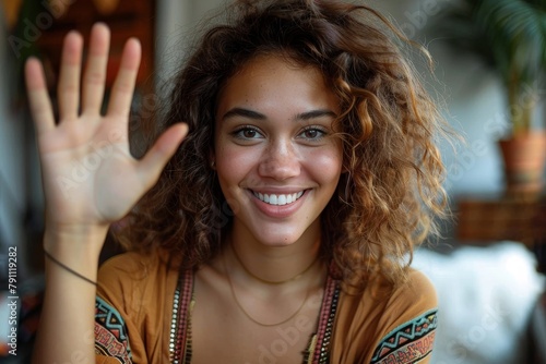 Smiling young woman with curly hair is waving at the camera with a friendly expression, in a natural indoor setting