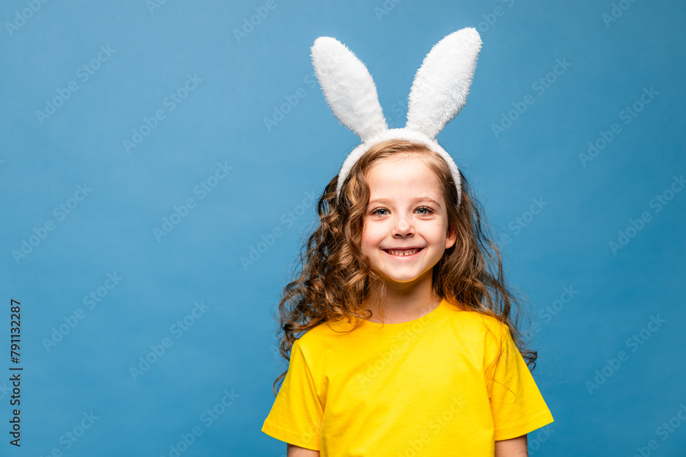 Cute little girl with curly hair in yellow t-shirt with bunny ears on blue background. Easter celebration concept